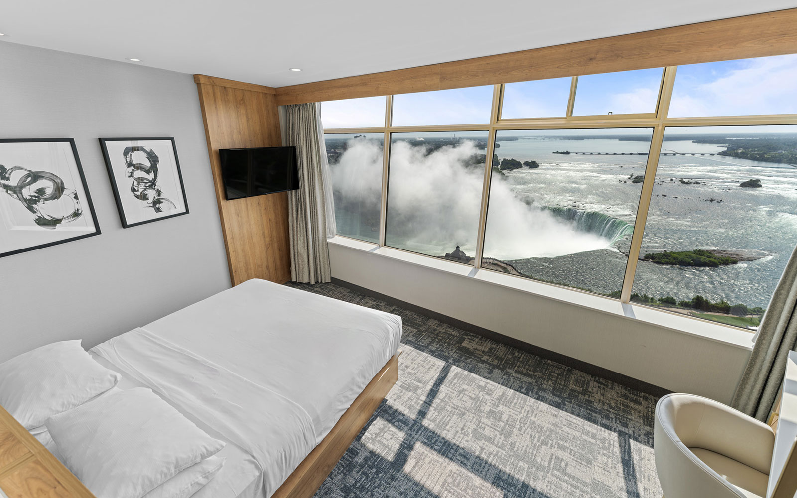 Hotel room with panoramic Niagara Falls views, white bed, light gray walls, abstract art, flat-screen TV, beige carpet, and a cream-colored chair, all designed for luxury and natural light.