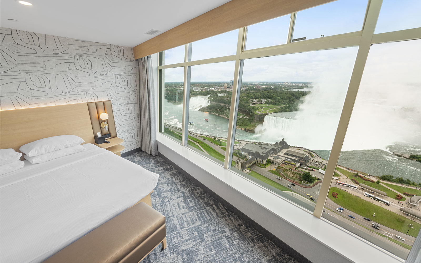 A modern hotel bedroom suite with a light beige and gray color scheme, featuring large windows offering a stunning view of Niagara Falls.