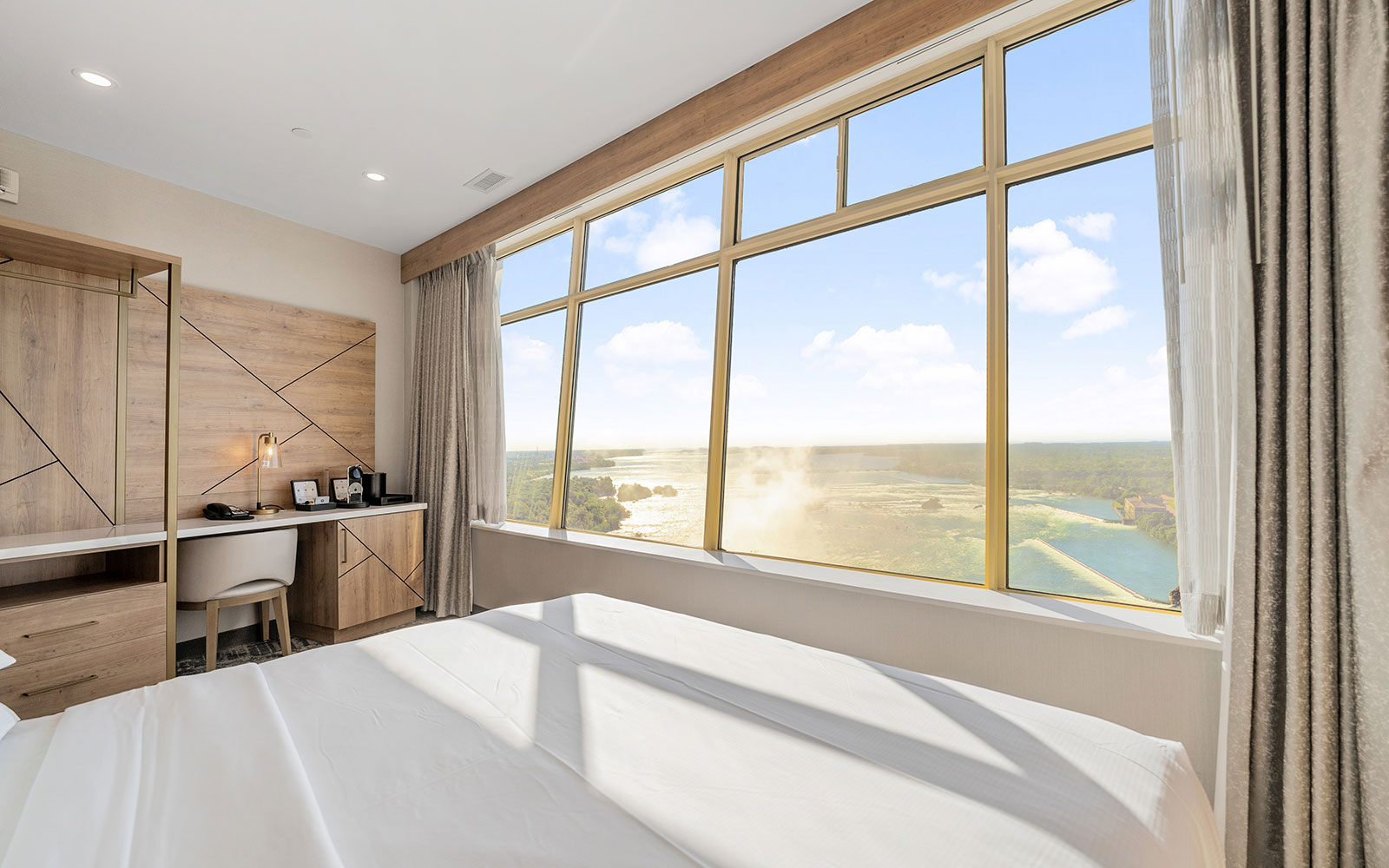 Hotel room with a large window offering a view of water, greeneries, and a pale sky, featuring a bed with white linens, light wooden headboard, small desk area, beige chair, light wooden wall unit, and soft natural lighting.